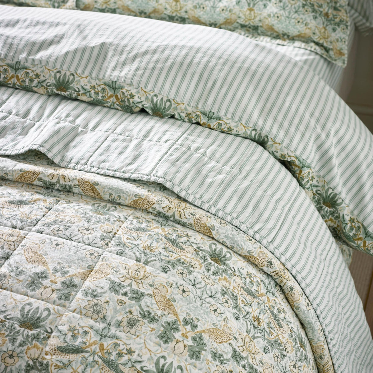 Strawberry Thief quilted cotton bedspread in sage green, showing bird and floral pattern alongside striped pillowcases with floral-trimmed edges on a made bed.