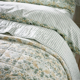 Strawberry Thief quilted cotton bedspread in sage green, showing bird and floral pattern alongside striped pillowcases with floral-trimmed edges on a made bed.