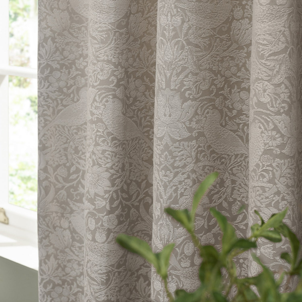 Pencil pleat curtains in linen tone showing the Strawberry Thief jacquard woven pattern with birds and foliage, pictured hanging beside a window with a green plant in view.