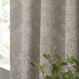 Pencil pleat curtains in linen tone showing the Strawberry Thief jacquard woven pattern with birds and foliage, pictured hanging beside a window with a green plant in view.