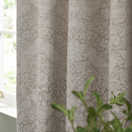 Pencil pleat curtains in linen tone showing the Strawberry Thief jacquard woven pattern with birds and foliage, pictured hanging beside a window with a green plant in view.