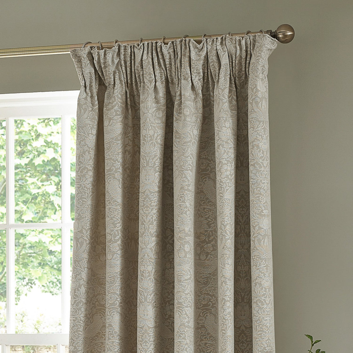 Strawberry Thief Jacquard Linen pencil pleat curtains in beige, shown hanging from a pole with pinch-pleat header, woven floral and bird pattern, in a room beside a window.
