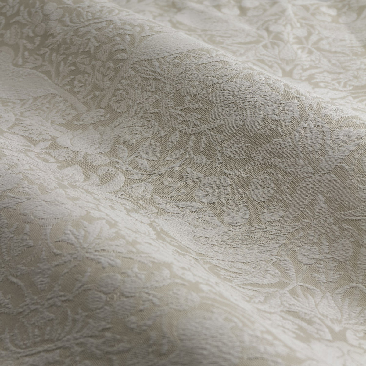Close-up of Strawberry Thief jacquard linen pencil pleat curtain fabric in beige, showing a woven tonal pattern of strawberry plants and birds catching light across soft folds.