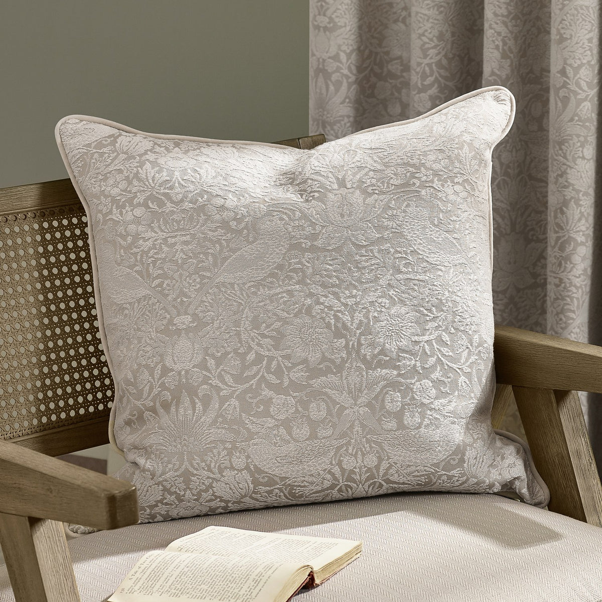 Strawberry Thief Jacquard Linen Cushion in cream, woven bird and strawberry motif on a linen-toned ground with piped edge, shown on a wooden armchair.