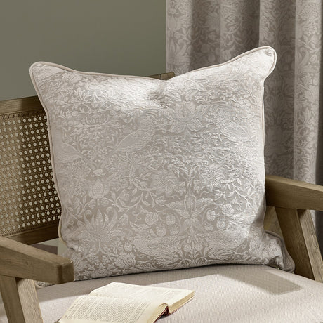 Strawberry Thief Jacquard Linen Cushion in cream, woven bird and strawberry motif on a linen-toned ground with piped edge, shown on a wooden armchair.
