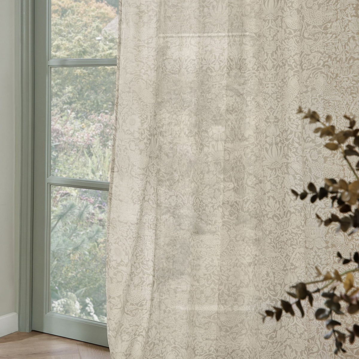 Strawberry Thief Floral Linen Curtain Panel in cream, semi-sheer fabric with tonal floral print, shown hanging beside a window overlooking greenery.