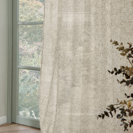 Strawberry Thief Floral Linen Curtain Panel in cream, semi-sheer fabric with tonal floral print, shown hanging beside a window overlooking greenery.