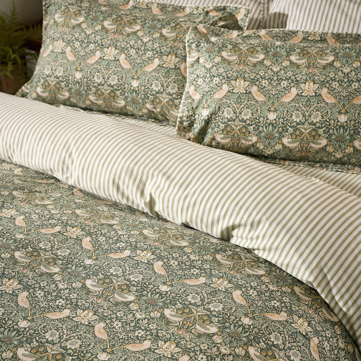 Strawberry Thief cotton duvet cover set in forest green, showing bird and floral pattern pillowcases paired with striped reverse on a made bed.