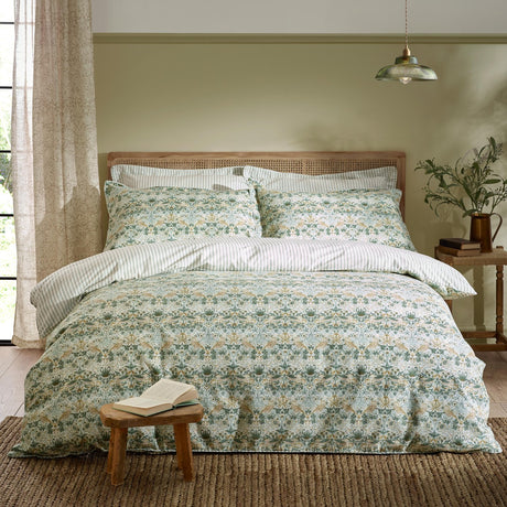 Strawberry Thief cotton floral duvet cover set in sage green with matching striped pillowcases, styled on a bed with a rattan headboard in a bedroom setting.