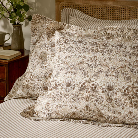 Strawberry Thief Percale Cotton Pillowcase Pair in Natural, styled on a bedroom pillow with bird and floral print, shown alongside striped bedding and a cane headboard.
