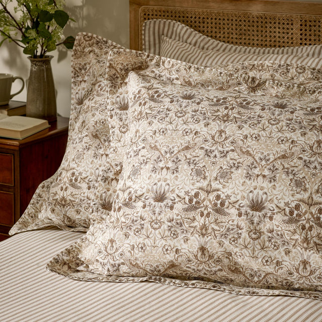 Strawberry Thief Percale Cotton Pillowcase Pair in Natural, styled on a bedroom pillow with bird and floral print, shown alongside striped bedding and a cane headboard.