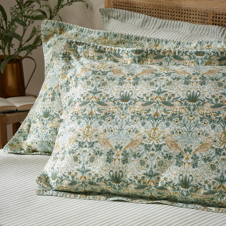 Strawberry Thief percale cotton pillowcase pair in sage green, styled on a bed with striped linen, showing the floral bird-and-foliage print in green, gold, and cream tones.