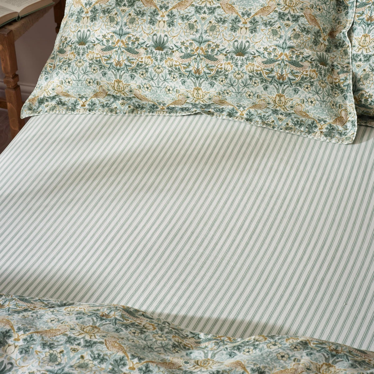 Strawberry Thief Percale Cotton Fitted Sheet in sage green, fine narrow stripe pattern, shown on a bed with matching floral pillowcases and duvet cover.