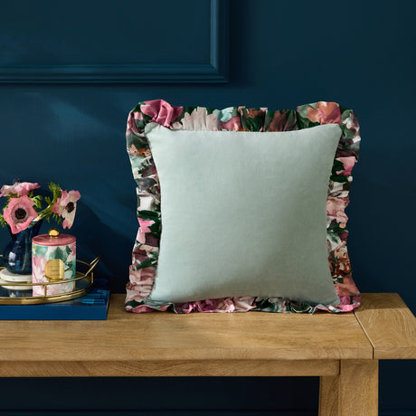 Tahiti Velvet Cotton Cushion in Duck Egg with a plain green velvet face and pink floral-print frill, shown on a wooden bench against a dark blue wall.
