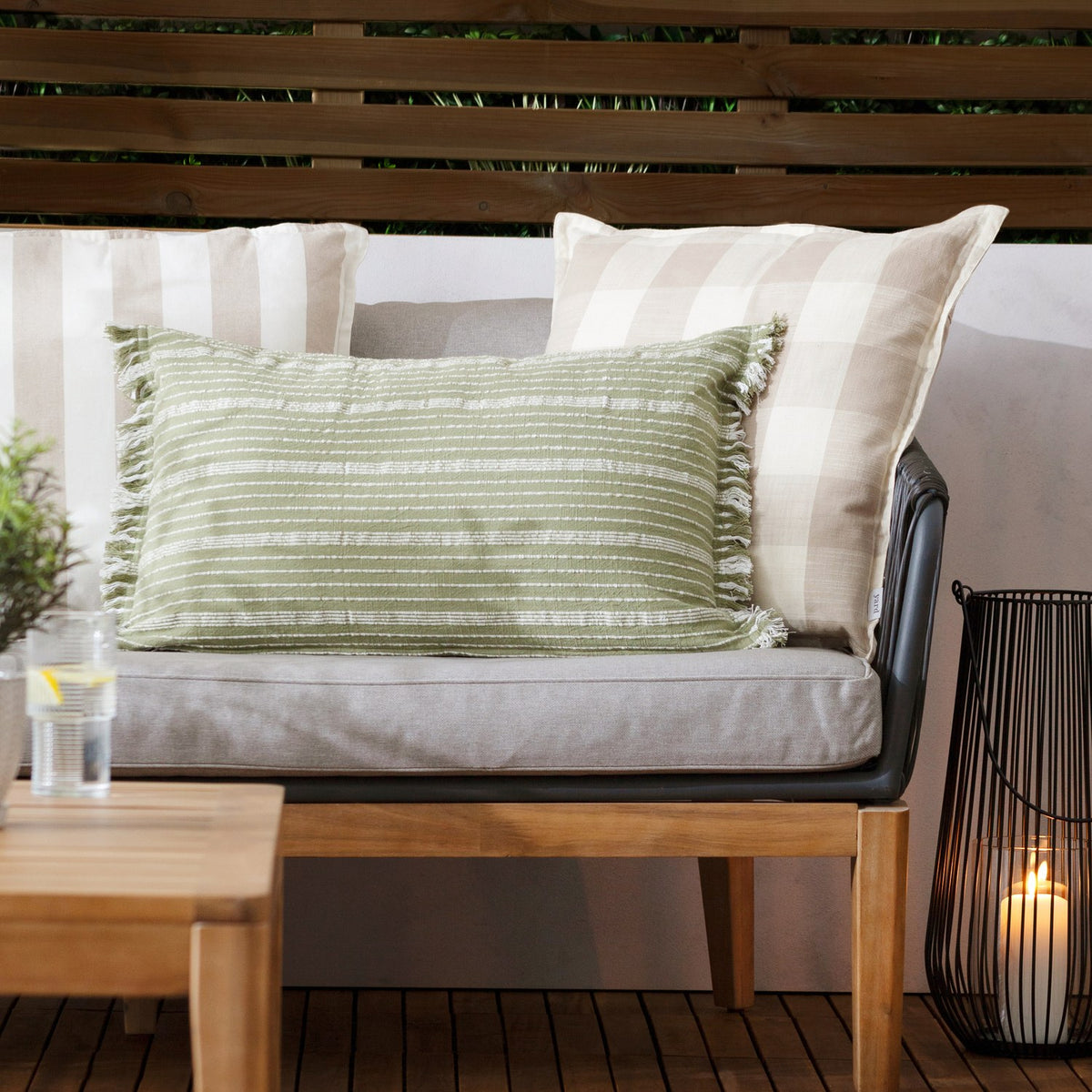 Tide Striped Cotton Cushion in moss green, a rectangular lumbar cushion with fringed edge trim, shown on an outdoor sofa alongside beige check and stripe scatter cushions.