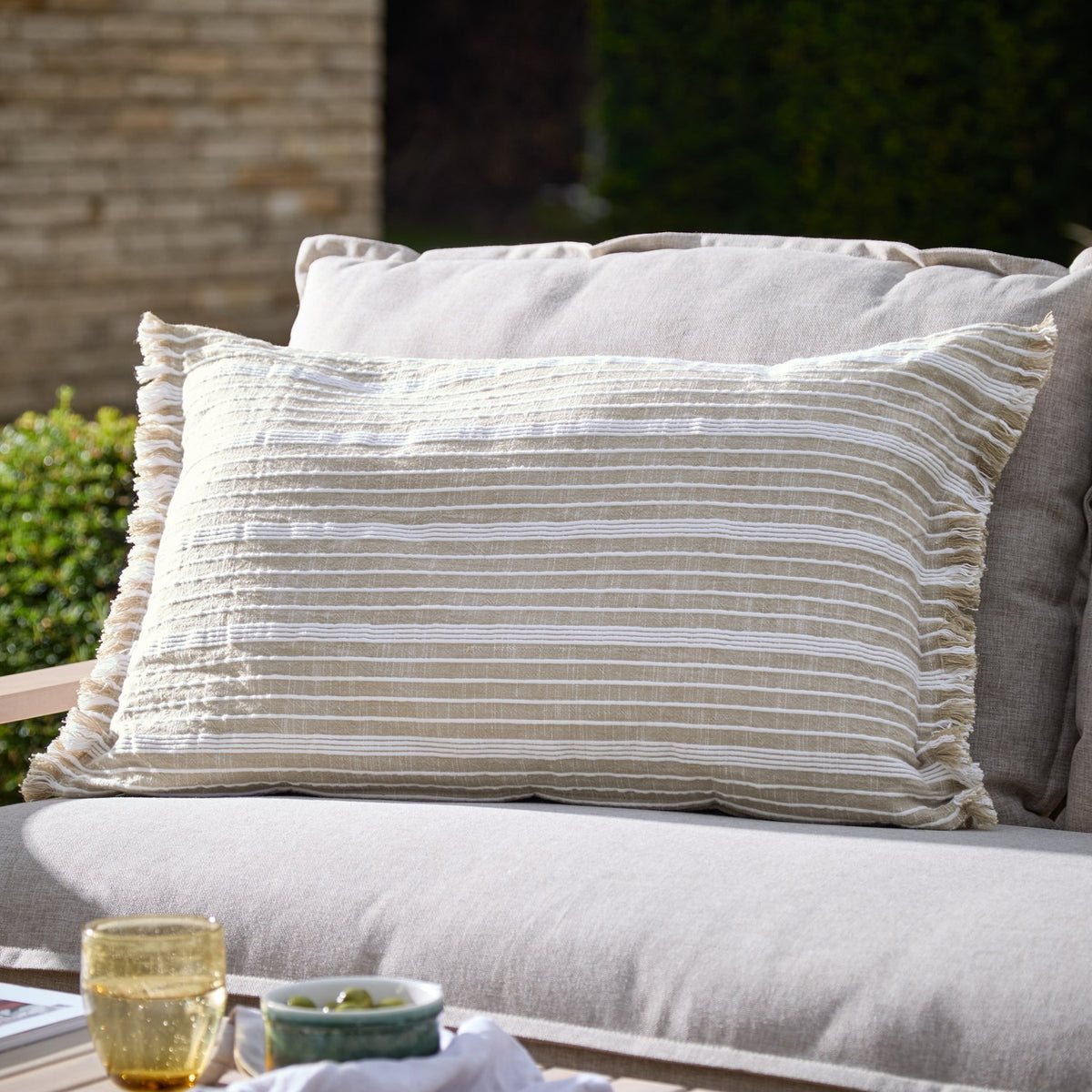 Tide Striped Cotton Cushion in natural beige with fine woven white stripes and a fringed edge, shown on an outdoor sofa in a garden setting.