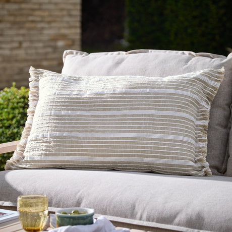 Tide Striped Cotton Cushion in natural beige with fine woven white stripes and a fringed edge, shown on an outdoor sofa in a garden setting.