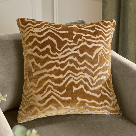 Tierra Abstract Velvet Cushion in brass gold, square with a wandering cut-velvet chevron pattern, shown on a grey armchair.