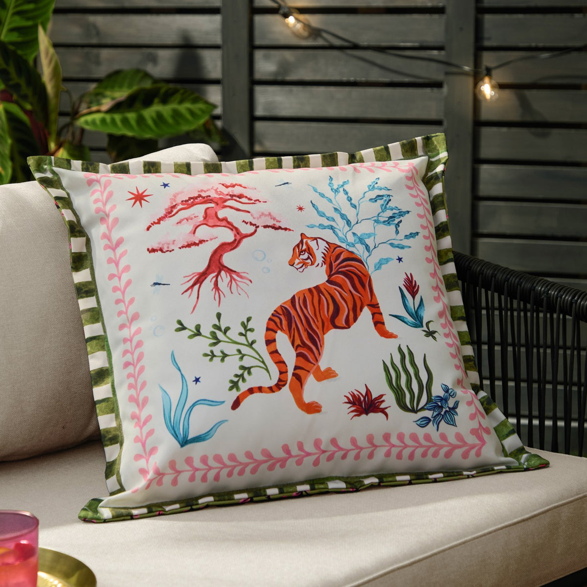 Tiger Dream reversible polyester cushion in cream on an outdoor sofa, featuring an orange tiger, coral branches and pink foliage border with green striped trim.