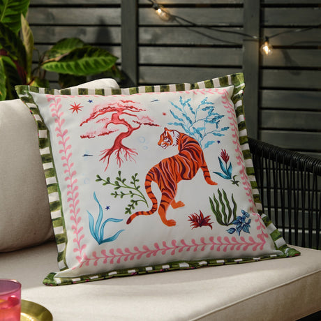 Tiger Dream reversible polyester cushion in cream on an outdoor sofa, featuring an orange tiger, coral branches and pink foliage border with green striped trim.