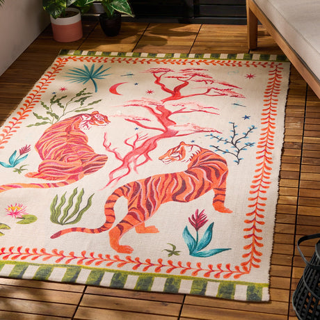Tiger Dream Jungle rug in multi polyester, printed with two orange tigers beside a pink tree on cream ground, framed by a green-and-cream striped border, shown on an outdoor wooden deck.