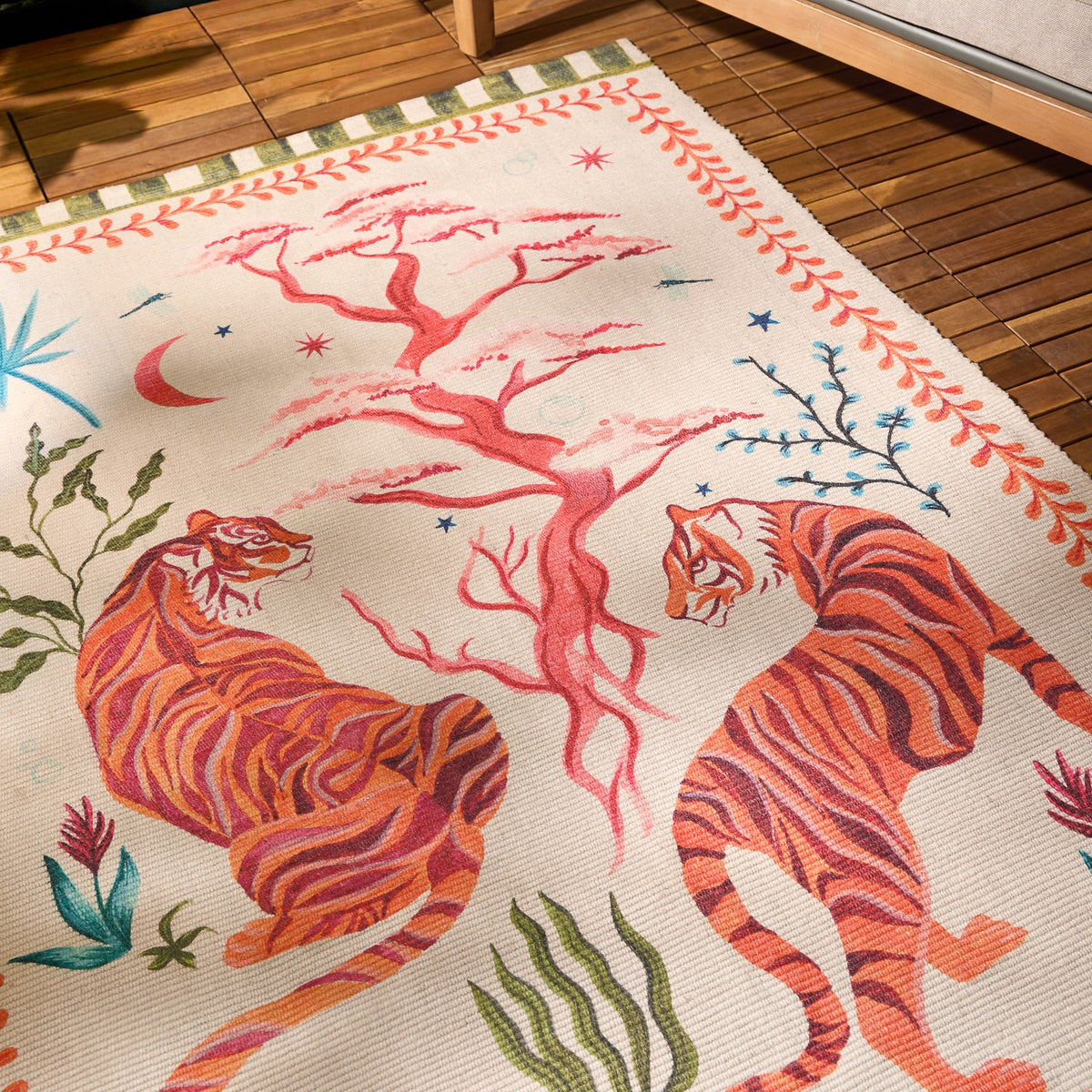 Tiger Dream Jungle Polyester Rug in Multi, cream ground with two orange tigers flanking a coral-pink tree, moon and stars motifs, green-striped border, shown on a wooden floor.