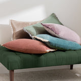 Tilly reversible velvet cushion in dusky pink, shown among coordinating green, mauve, blue and shell-toned bolster cushions with ruffle-edge trims on a green upholstered bench.