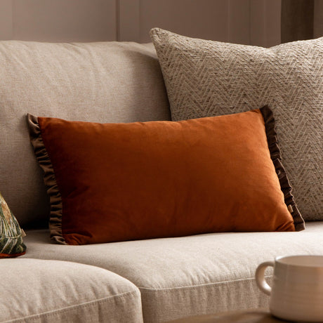 Tilly reversible rectangular velvet cushion in rust with a frilled edge trim, shown on a beige sofa alongside a herringbone cushion.