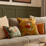 Rust velvet cushion with frilled edge trim shown on a sofa alongside mushroom-print and mustard embroidered cushions in a living room setting.