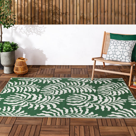 Tocorico Jacquard Jungle Rug in green and white, rectangular with a leaf print, shown on a wooden deck beside a chair and potted plants.