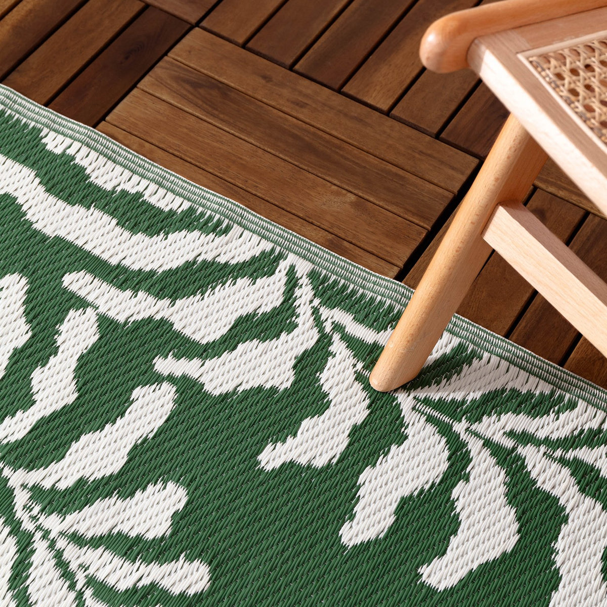 Tocorico Jacquard Jungle Rug in green and white with a leaf pattern, shown on a wooden deck beneath a light wood chair leg.