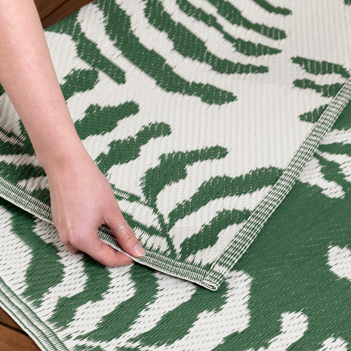 Hand folding back the corner of the Tocorico Jacquard Jungle Rug, showing a green and white leaf pattern woven into the polypropylene outdoor rug's edge and underside.
