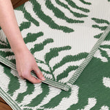Hand folding back the corner of the Tocorico Jacquard Jungle Rug, showing a green and white leaf pattern woven into the polypropylene outdoor rug's edge and underside.