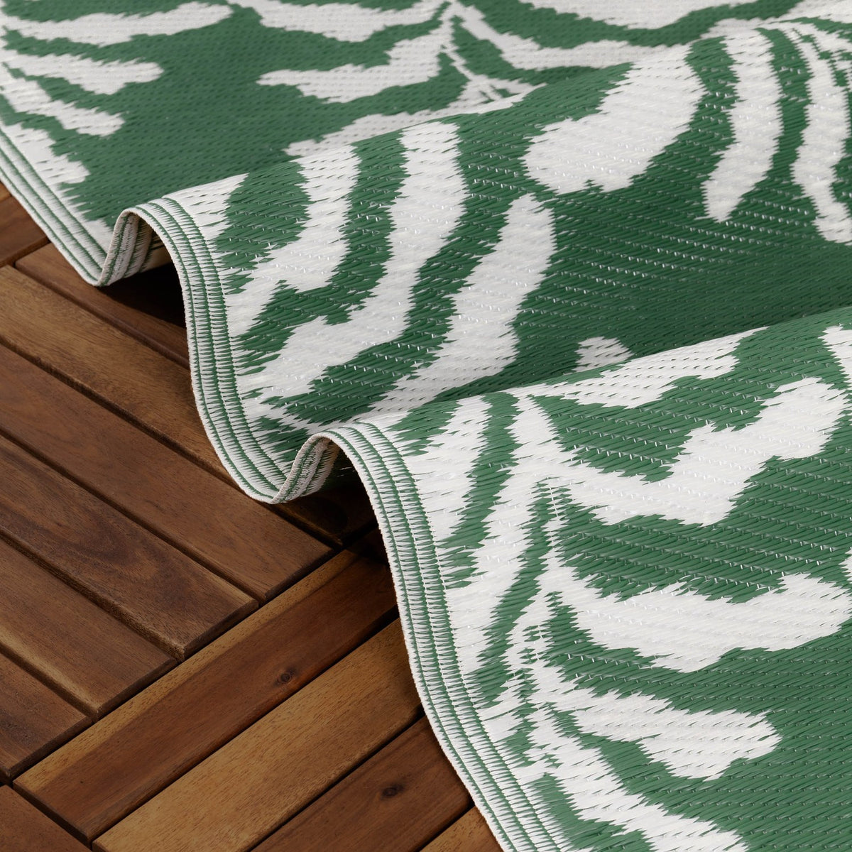 Tocorico Jacquard Jungle Rug in green and white, folded to show layered edges with a leaf print, laid over a wooden deck outdoors.