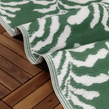 Tocorico Jacquard Jungle Rug in green and white, folded to show layered edges with a leaf print, laid over a wooden deck outdoors.