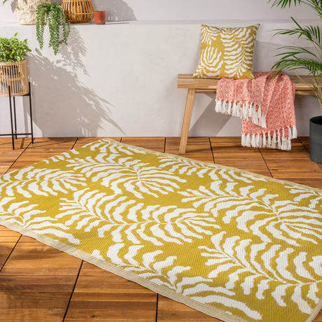 Tocorico Jacquard Jungle Rug in mustard yellow and white, woven with a fern leaf pattern, shown flat on wooden decking beside a bench with cushion and throw.
