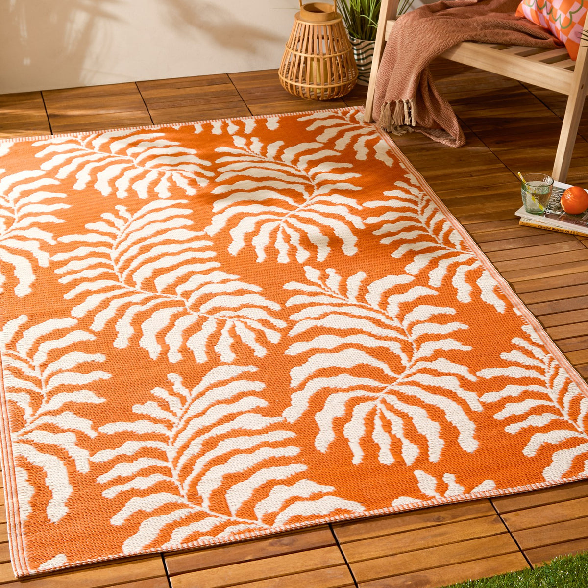 Tocorico Jacquard Jungle Rug in orange and cream, woven leaf pattern, shown on a wooden deck beside a chair and lantern in an outdoor setting.