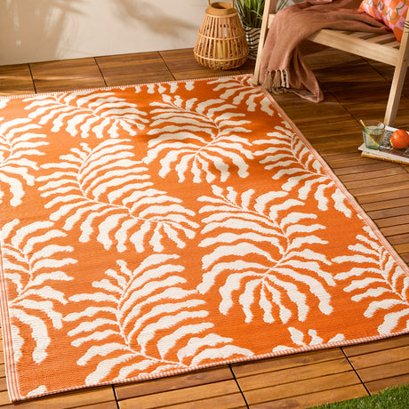 Tocorico Jacquard Jungle Rug in orange and cream, woven leaf pattern, shown on a wooden deck beside a chair and lantern in an outdoor setting.