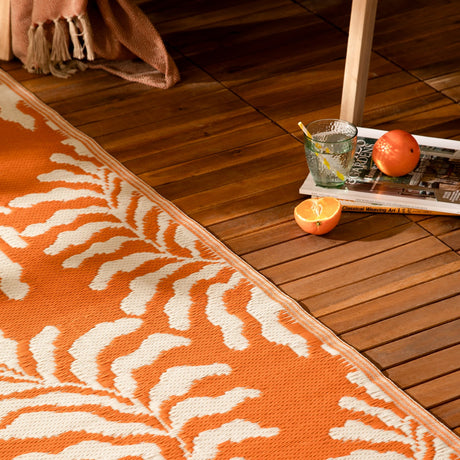Tocorico Jacquard Jungle Rug in orange and cream, textured leaf pattern, shown on wooden decking beside citrus fruit and a magazine.