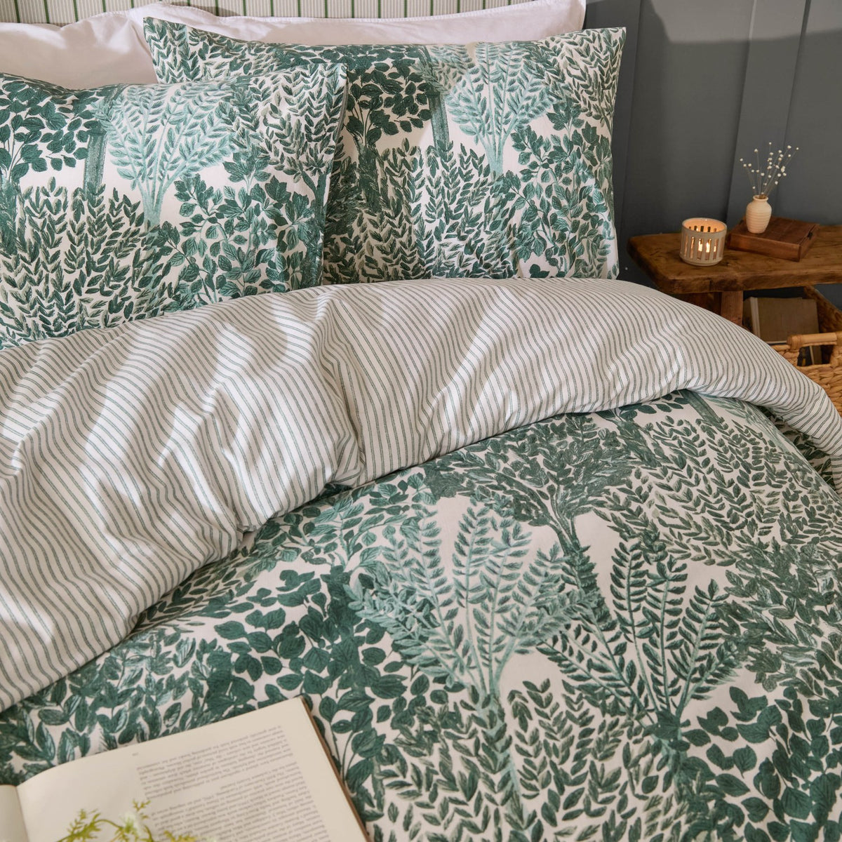Green Topiary Reversible Botanical Duvet Cover Set with matching pillowcases on a bed, leaf print facing up and striped reverse peeking through, in a bedroom with a bedside table and candle.