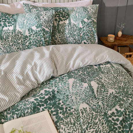 Green Topiary Reversible Botanical Duvet Cover Set with matching pillowcases on a bed, leaf print facing up and striped reverse peeking through, in a bedroom with a bedside table and candle.