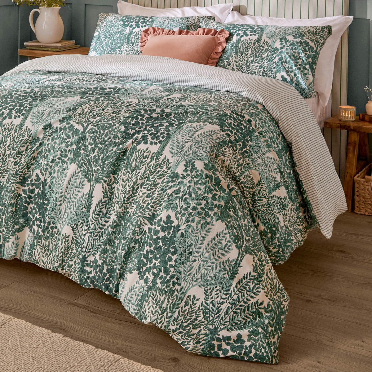 Green Topiary Reversible Botanical Duvet Cover Set with fern print on a bedroom bed, styled with matching pillowcases and a pink cushion, striped reverse edge visible.