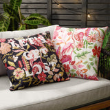 Tropical Bloom Lost reversible polyester cushion in black with coral, pink and olive floral print and "Let's Get Lost" text, shown on an outdoor sofa beside a matching cream floral cushion.