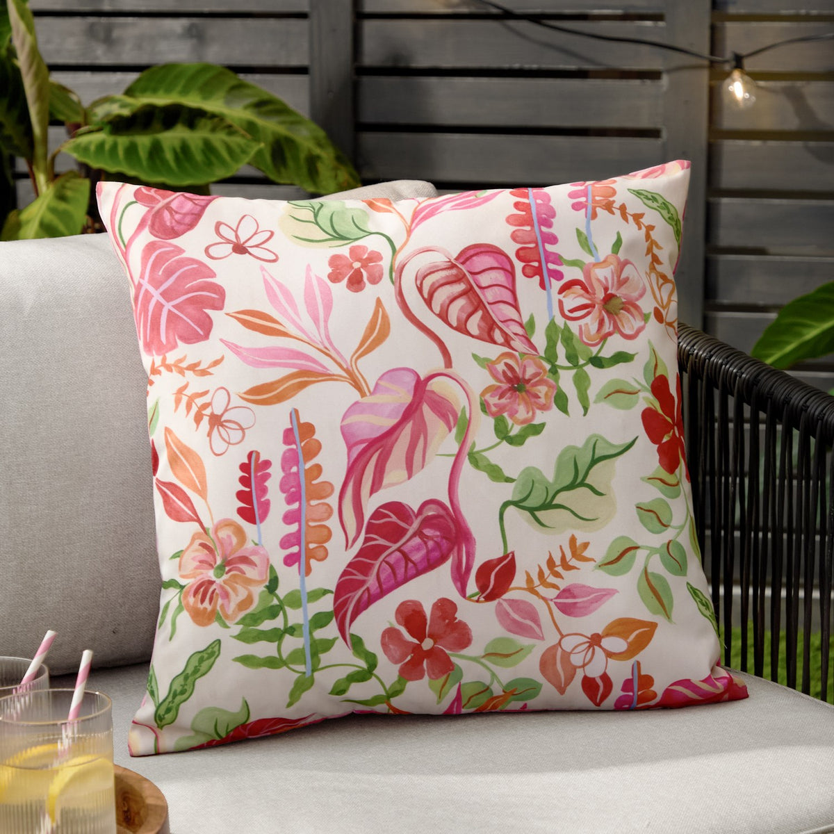 Tropical Bloom Reversible Polyester Cushion in natural cream, featuring a hand-illustrated tropical leaf and floral print in pink, coral and green, shown on an outdoor sofa.