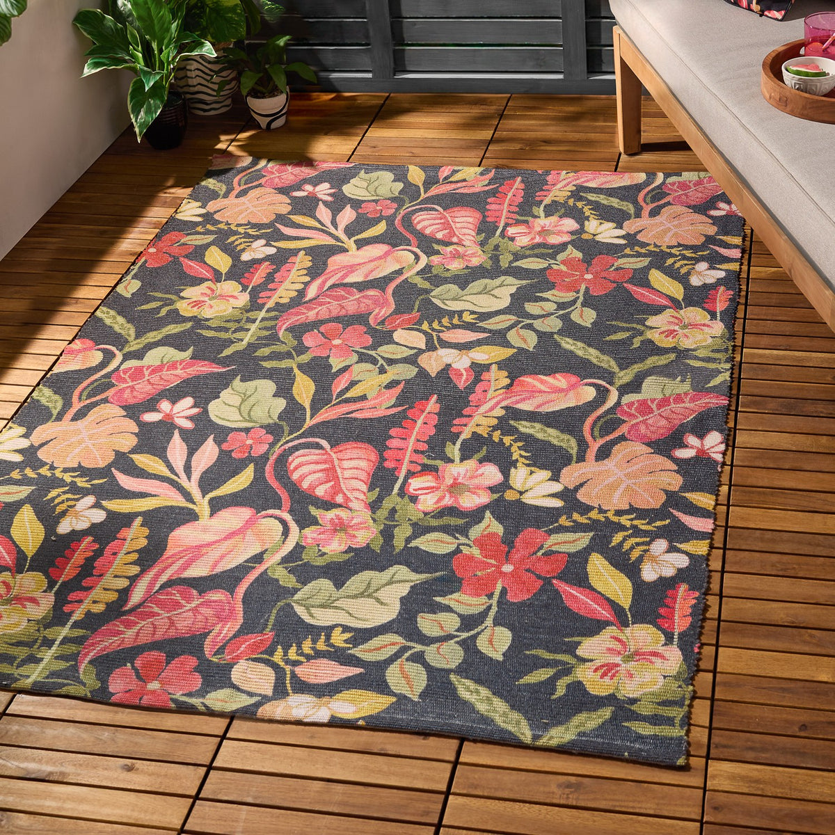 Tropical Bloom rectangular rug in polyester with a charcoal-black ground and dense tropical floral print in coral, blush, apricot and green, shown on a wooden deck beside a sofa and potted plants.