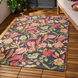 Tropical Bloom rectangular rug in polyester with a charcoal-black ground and dense tropical floral print in coral, blush, apricot and green, shown on a wooden deck beside a sofa and potted plants.
