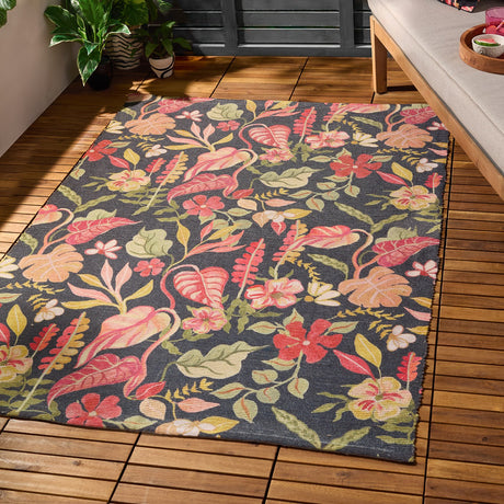 Tropical Bloom rectangular rug in polyester with a charcoal-black ground and dense tropical floral print in coral, blush, apricot and green, shown on a wooden deck beside a sofa and potted plants.