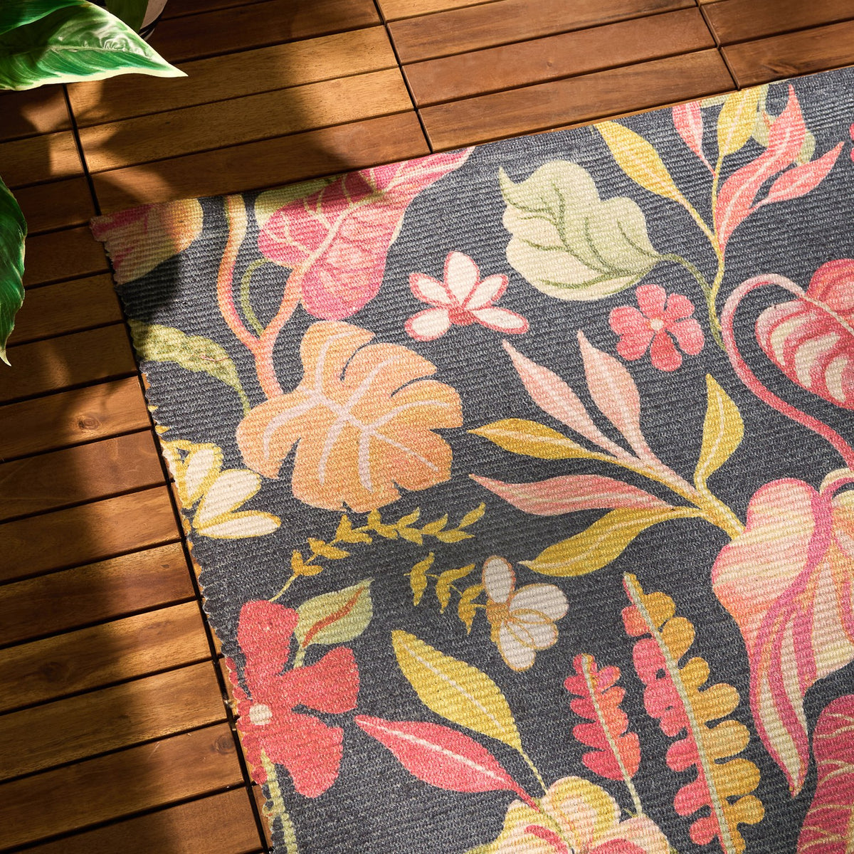 Tropical Bloom Floral Polyester Rug in black, corner view showing coral, pink and mustard leaf and floral prints on a charcoal ground, laid on a wooden deck.