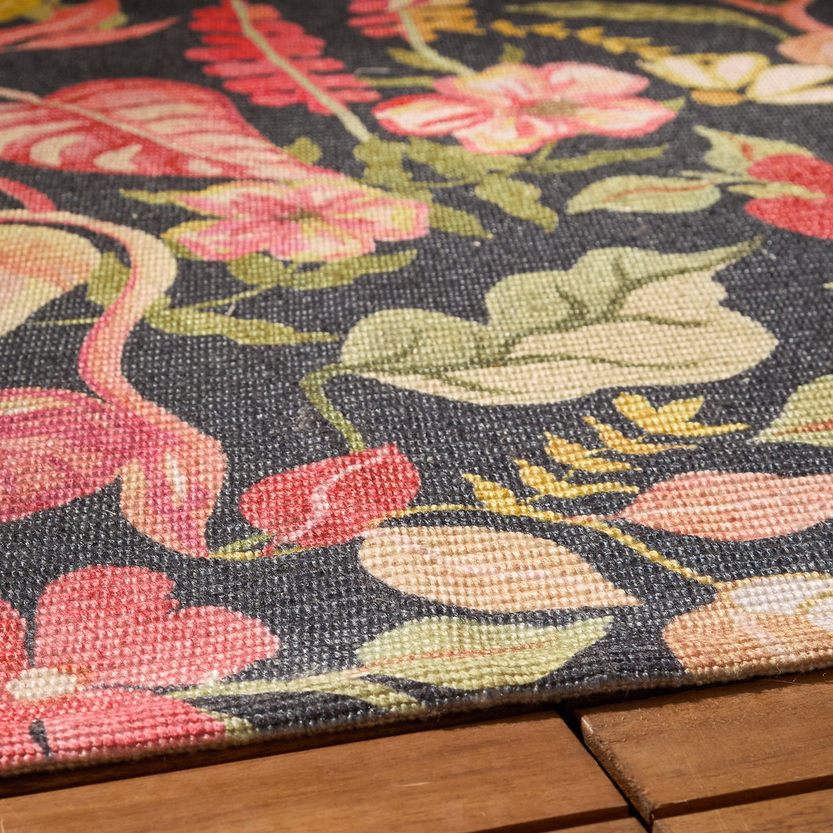 Close-up of the Tropical Bloom polyester rug's woven texture on a black ground, showing coral and blush leaves, apricot blooms and green foliage, corner edge resting on a wooden surface.