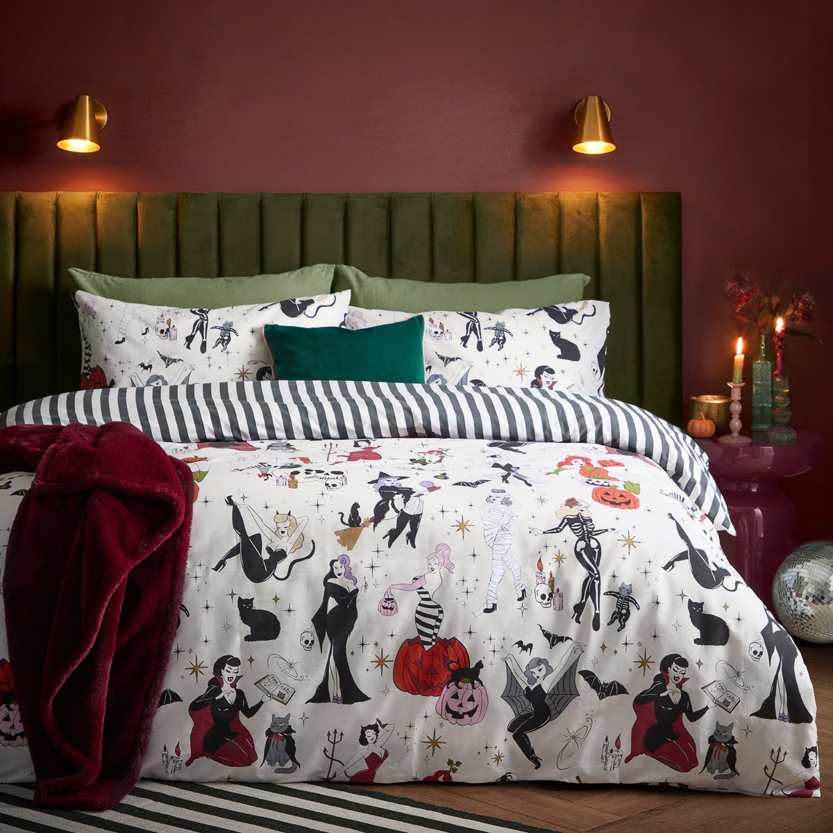 Trick or Treat reversible duvet cover set on a bed with Halloween-print witches, skeletons and pumpkins, paired with black and white striped pillowcases in a dark red bedroom.